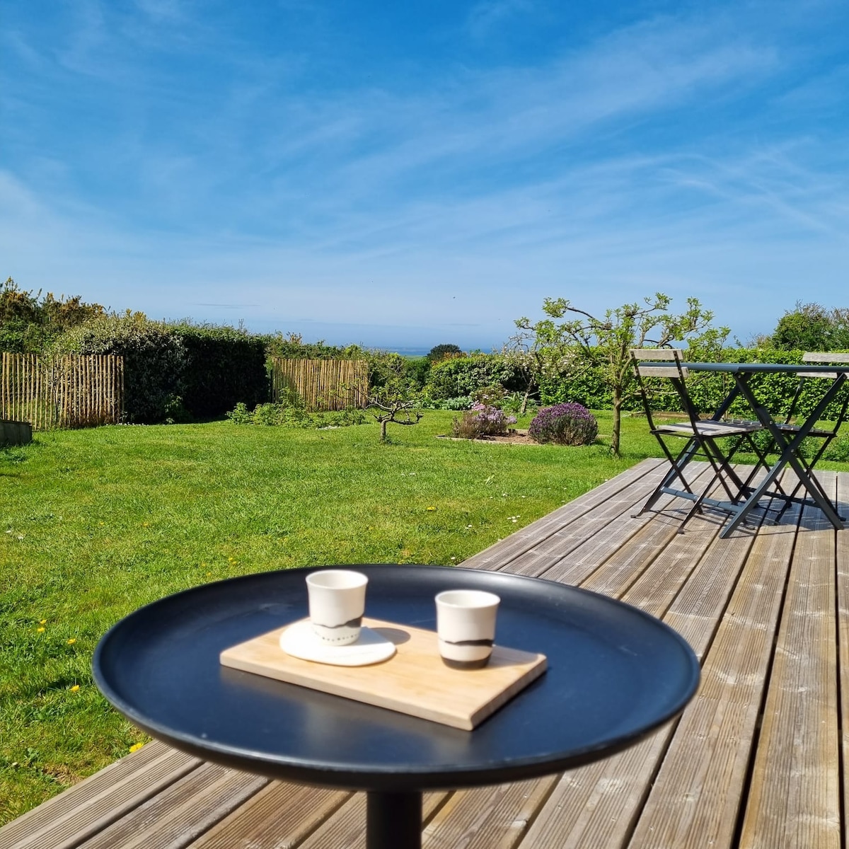A wooden deck extends into a lush garden, featuring green grass and blooming flowers. A small black table holds two coffee cups on a wooden tray, while a set of metal chairs is visible nearby, offering a space for relaxation and enjoying the outdoor view.