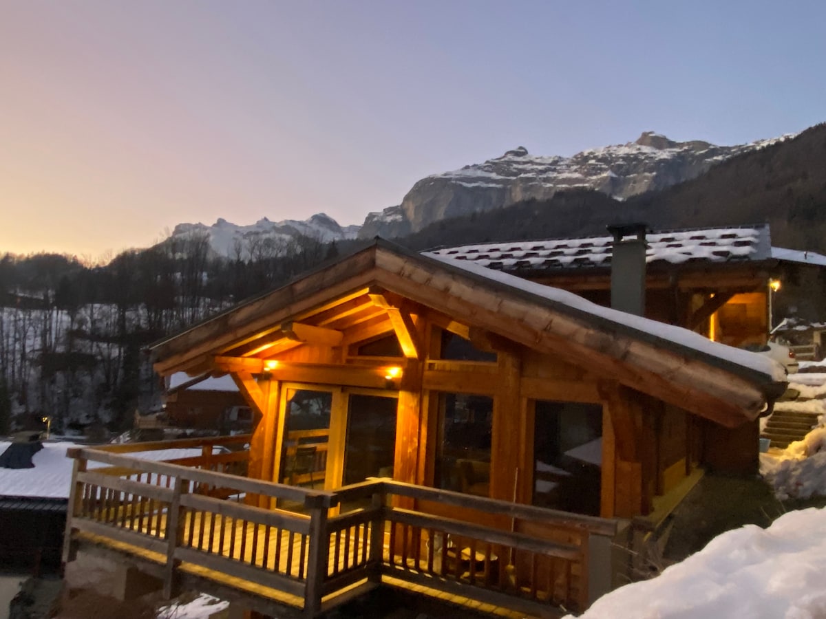 A wooden chalet is illuminated by warm outdoor lighting, showcasing its rustic architecture against a backdrop of snow-covered mountains. Large glass doors offer a glimpse into the cozy interior, while a wooden balcony provides access to the scenic landscape.