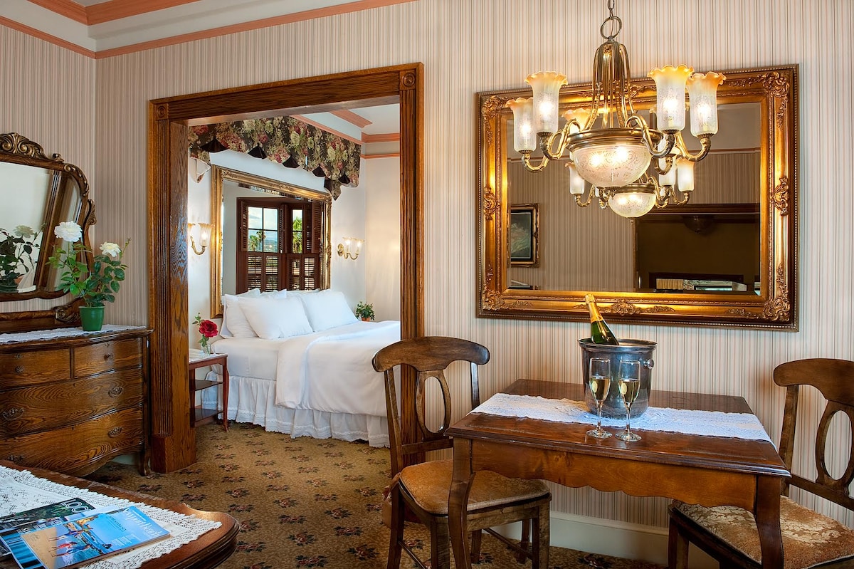 A Victorian-style room with wooden accents features a dining area with a table and two chairs. A large mirror reflects soft lighting from a chandelier above. The bedroom area, seen through an archway, contains a spacious bed and a decorative headboard, enhancing the room's elegant charm.
