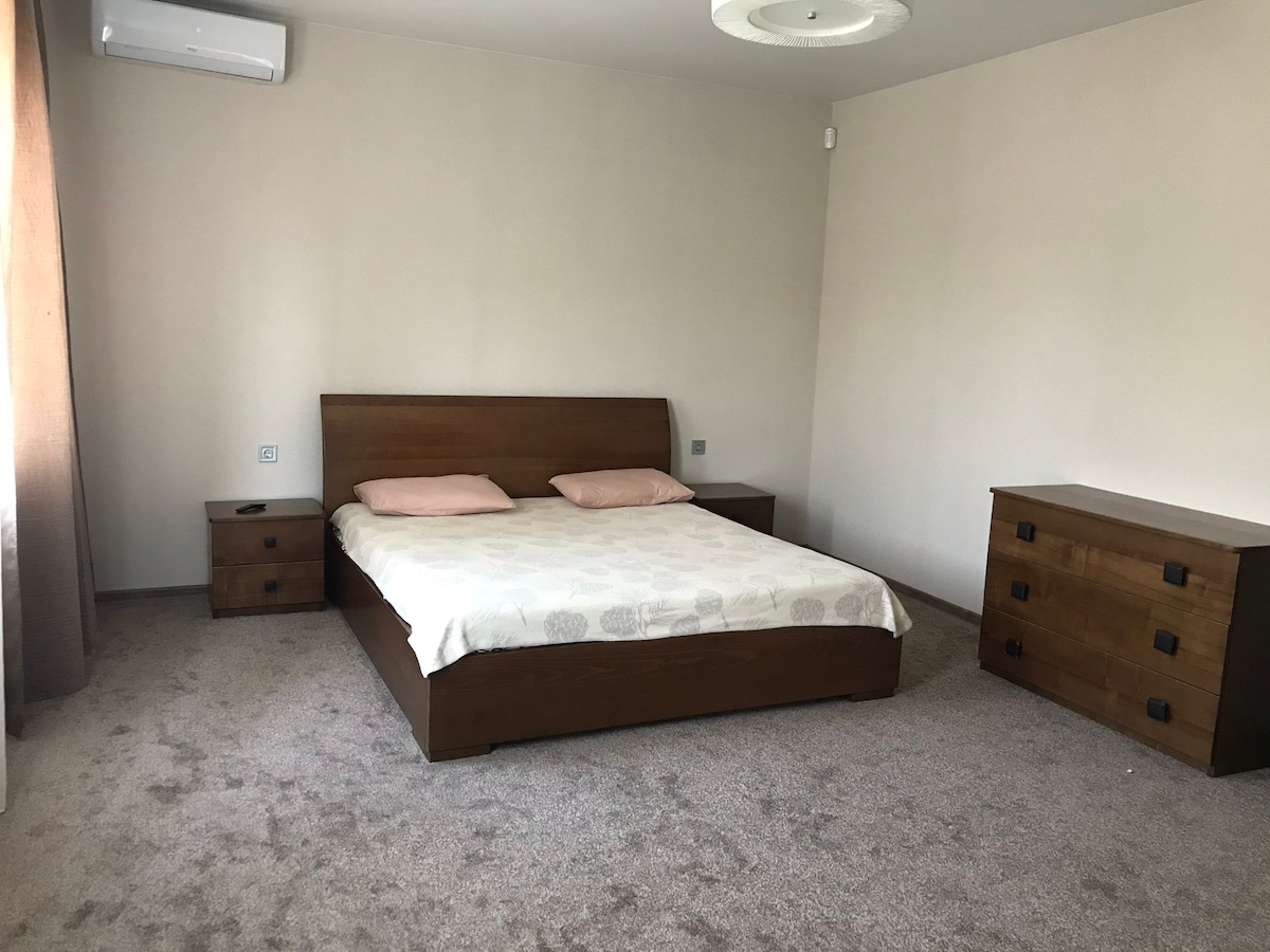 A spacious bedroom features a wooden bed with light-toned bedding and two nightstands. A matching dresser is positioned against the wall. Soft lighting from a ceiling fixture enhances the neutral color scheme. Windows are draped with light curtains, allowing natural light to fill the room.