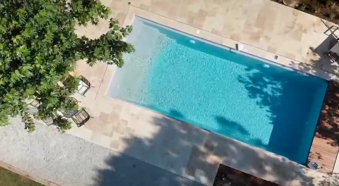 An aerial view of a rectangular heated pool, surrounded by natural stone decking. Sunlight reflects off the clear water, creating a tranquil atmosphere. Two chairs are positioned nearby, and greenery casts gentle shadows over the area, enhancing the sense of privacy.