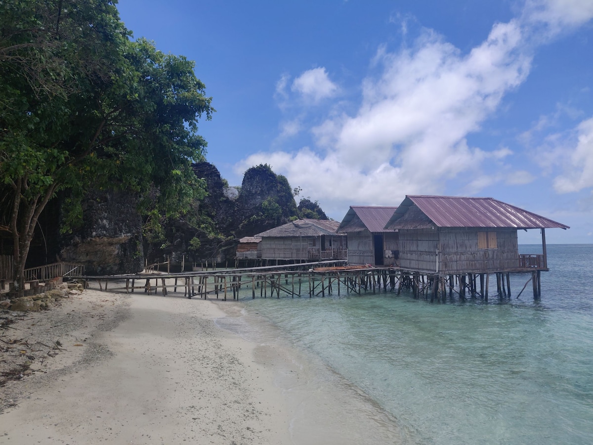 Two wooden structures are situated on stilts over calm water, connected by a wooden walkway. A sandy beach extends in front, with gentle waves lapping at the shore. Lush greenery frames the scene, and rocky outcrops are visible in the background under a partly cloudy sky.