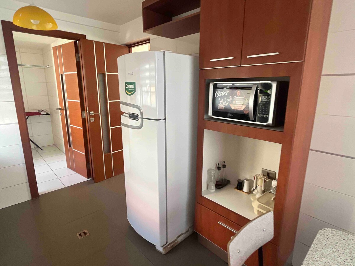 A modern kitchen space is depicted, featuring a stainless steel refrigerator and a microwave built into the cabinetry. Wooden cabinets provide storage, and a granite countertop is available for meal preparation. Light tiles cover the floor, contributing to a bright atmosphere.
