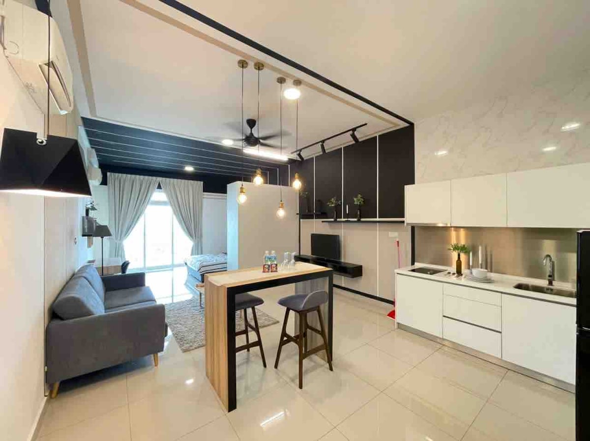 The modern living space features a comfortable gray sofa, a dining table with two stools, and a fully equipped kitchen. Warm lighting is provided by pendant lamps, while large windows allow natural light to fill the area. A cozy bedroom is visible in the background.