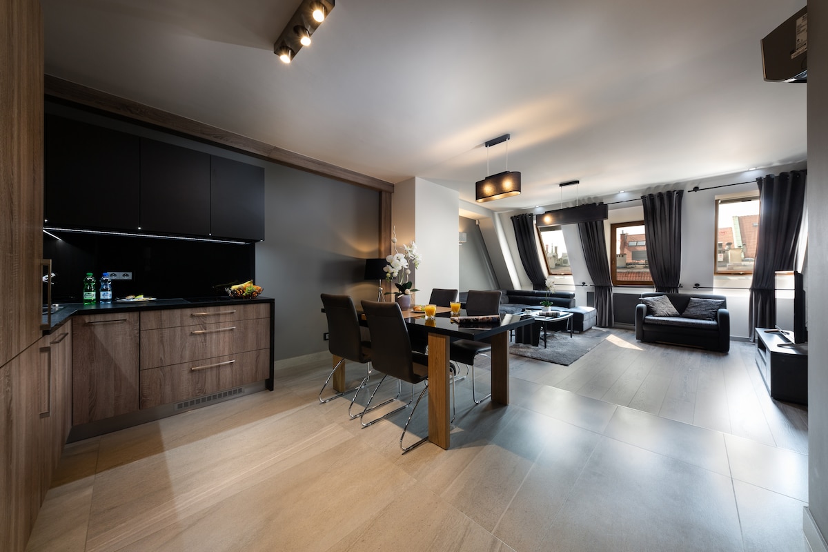 The open living area features a dining table with dark chairs and a kitchen with modern cabinetry. A spacious seating area is visible with a dark couch and a coffee table. Large windows allow natural light, and elegant drapes frame the view.