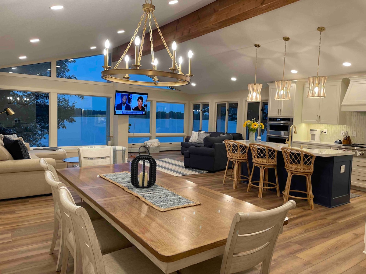 An open-plan living area features large windows overlooking the lake, bathed in evening light. A spacious dining table sits in the center, surrounded by stylish chairs. A comfortable seating area includes a sofa facing a wall-mounted television, with bar stools lining the kitchen island.