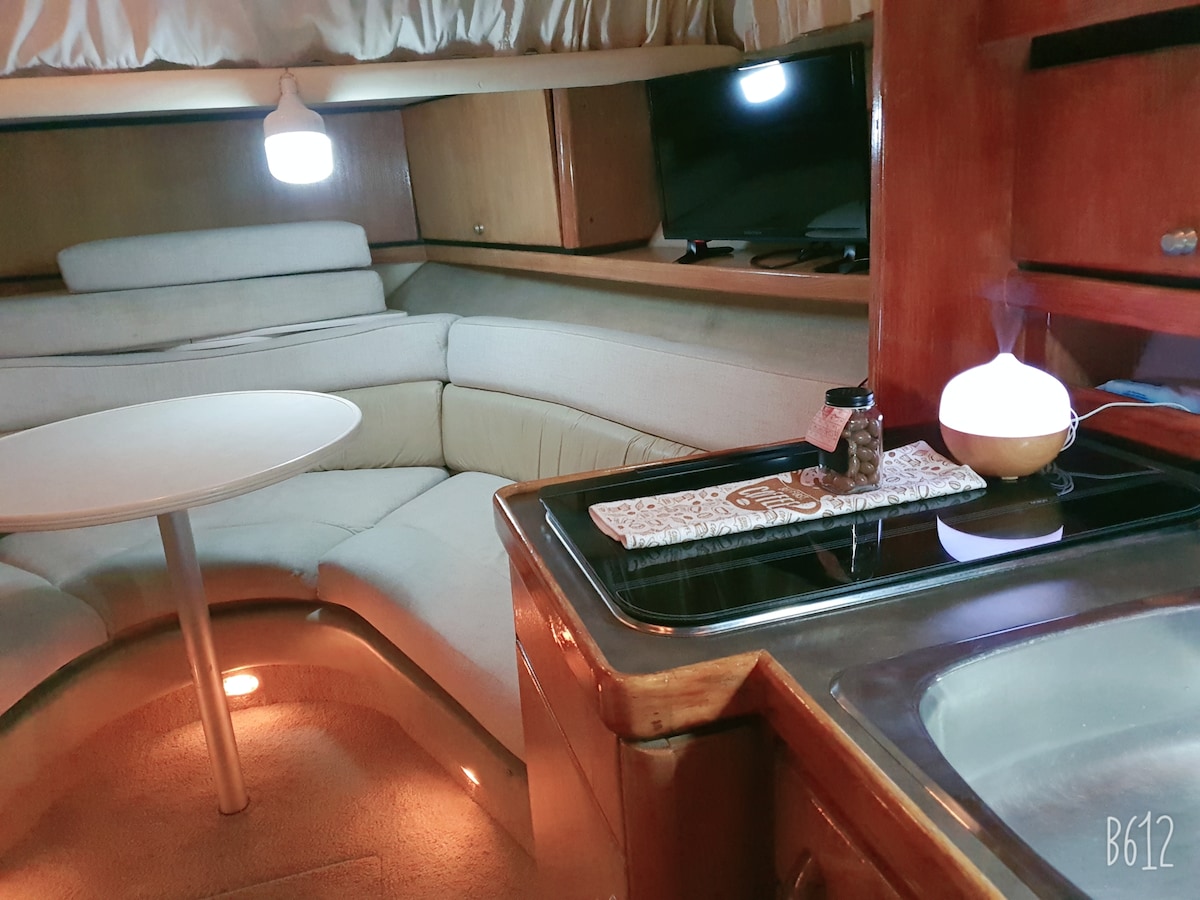An inviting interior space features a curved sofa surrounding a round table, with soft lighting from two lamps. A compact kitchen area showcases a stove and sink, enhancing functionality. Textures and finishes create a warm atmosphere, suggesting comfort and relaxation.