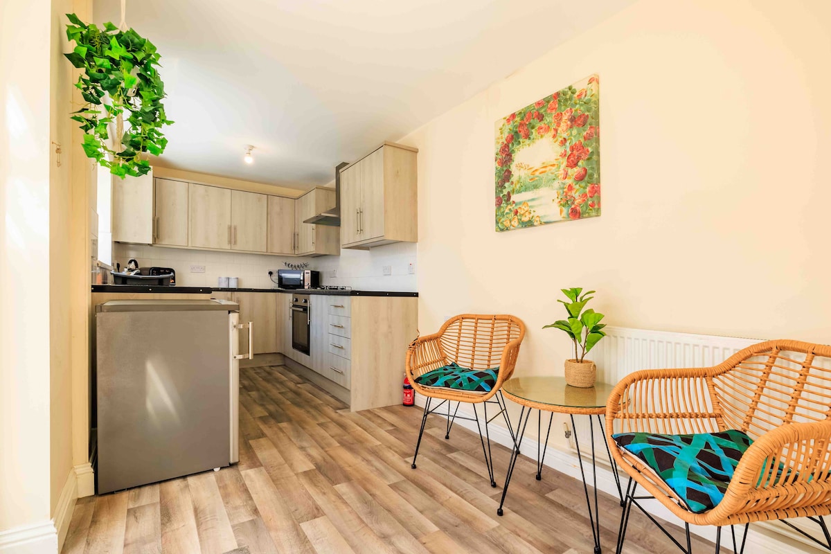 A newly refurbished kitchen area is shown, featuring modern appliances and light wood cabinetry. A small dining table is accompanied by two wicker chairs, each adorned with blue-green cushions. Natural light enhances the space, complemented by a wall-mounted floral artwork.
