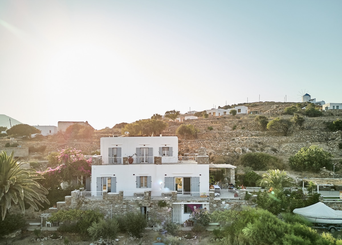 The exterior of the maisonette is presented, featuring a white facade complemented by surrounding greenery and stone pathways. The sun casts a warm glow over the structure, highlighting its spacious terraces and multiple balconies, offering views of the nearby hills.