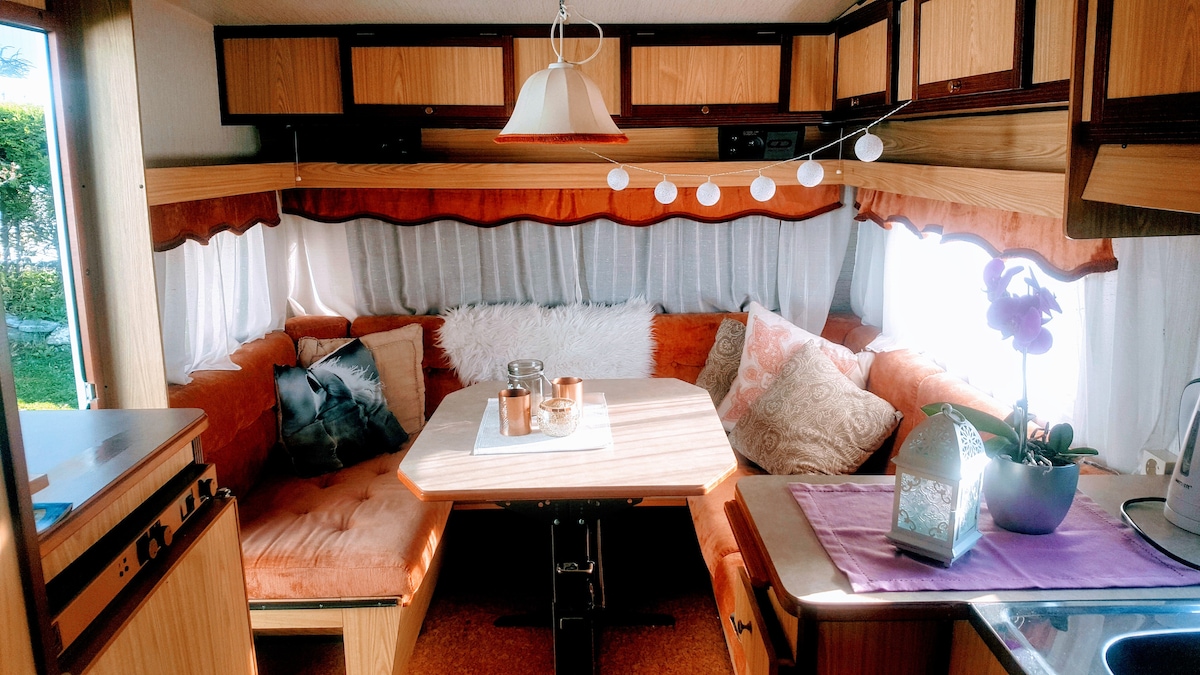A comfortable seating area is highlighted by vintage wood accents and soft cushions. A round table sits centrally, surrounded by a cozy orange leatherette seating. Natural light filters through sheer curtains, creating a warm atmosphere. Decorative items include a small plant, candles, and ambient lighting.