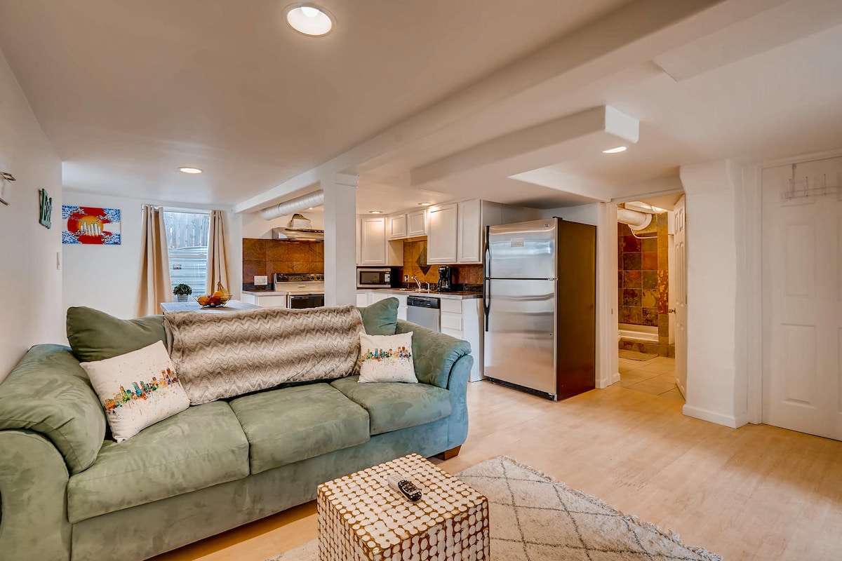 A cozy living area features a soft couch with decorative pillows, facing a coffee table. The space is illuminated by recessed lighting and natural light from nearby windows. A well-equipped kitchen area with modern appliances, including a refrigerator and stove, is observed in the background.