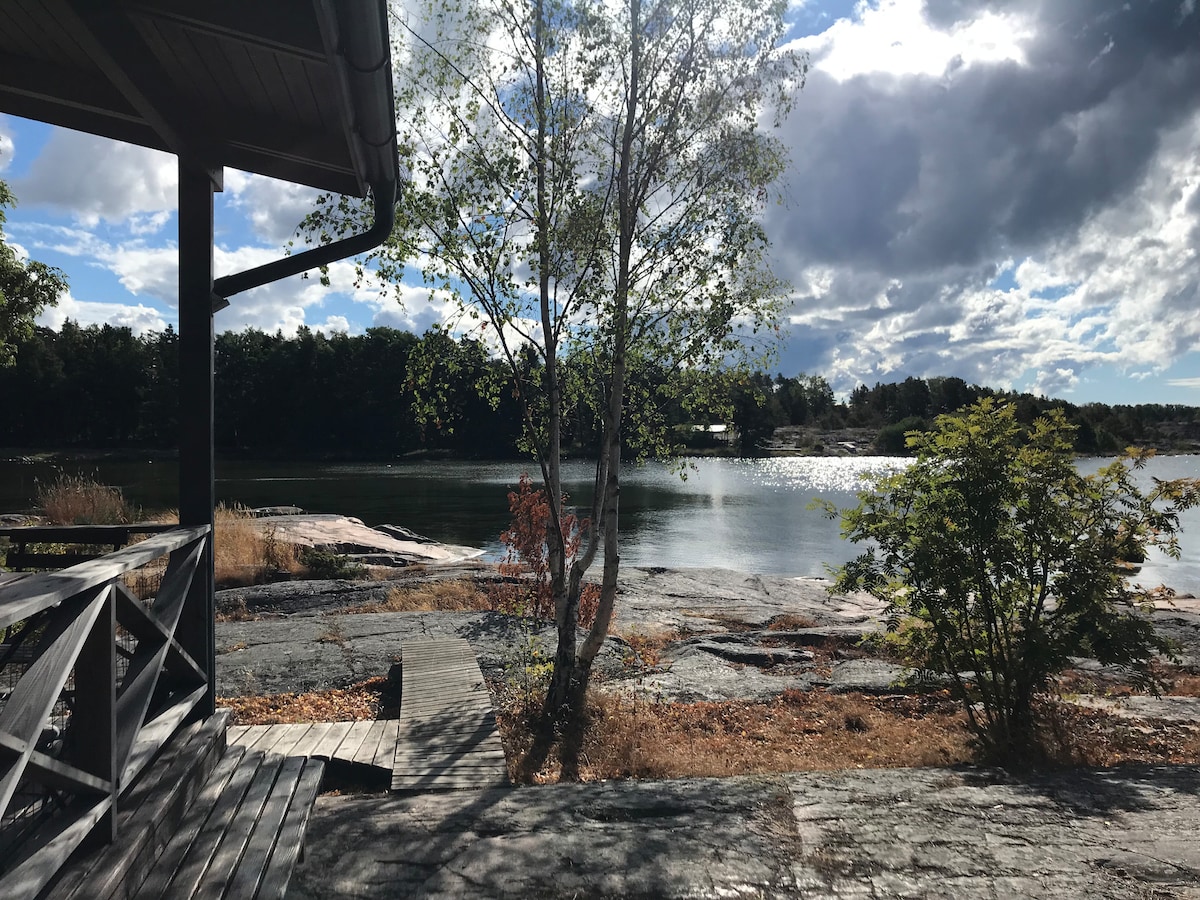A serene view of the water framed by a tree and rocks, with gentle sunlight reflecting on the surface. Seating is available on a wooden deck, providing a comfortable space to enjoy the peaceful surroundings.