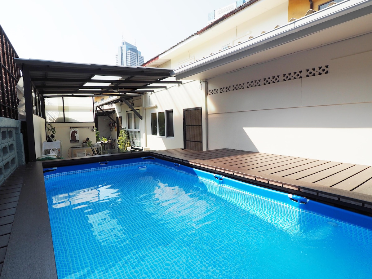 A private pool area is visible, featuring a well-maintained blue pool surrounded by wooden decking. A shaded seating area is positioned nearby, providing a relaxing space for guests. The background showcases part of the house with large windows allowing natural light.