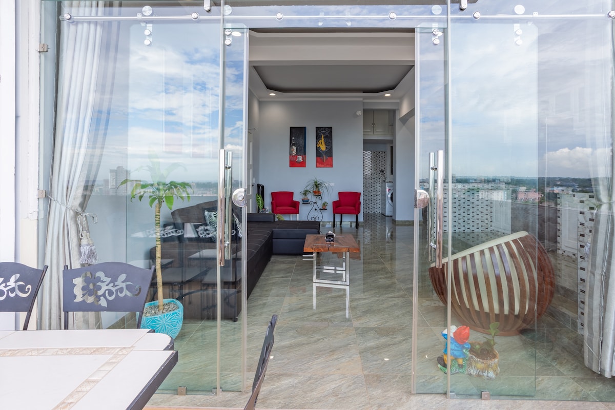 A spacious living area is visible through large glass doors, revealing modern furnishings in neutral tones. Two red chairs provide a pop of color, contrasted by a dark sofa and a decorative drum. Natural light floods the room, enhancing the open feel.
