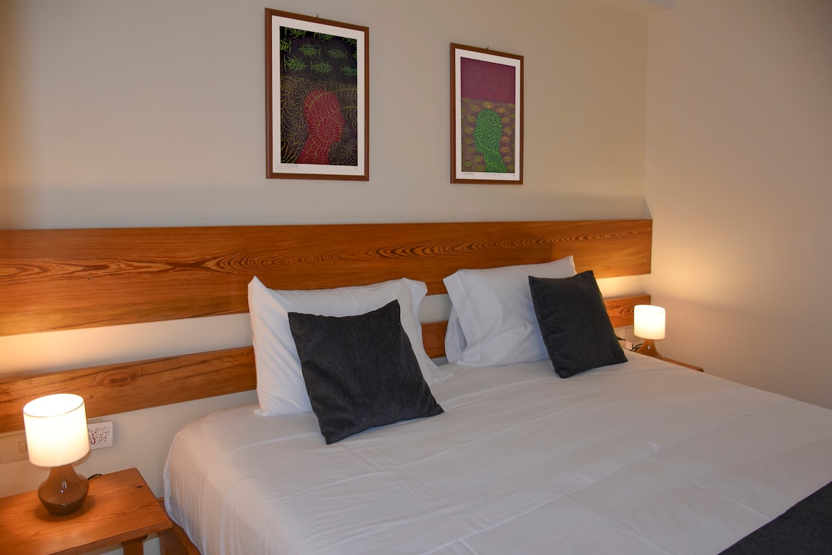 A spacious bed is positioned against a wooden headboard, complemented by two decorative pillows. Wall art featuring abstract designs hangs above. Soft lighting is provided by two bedside lamps, creating a calm atmosphere.