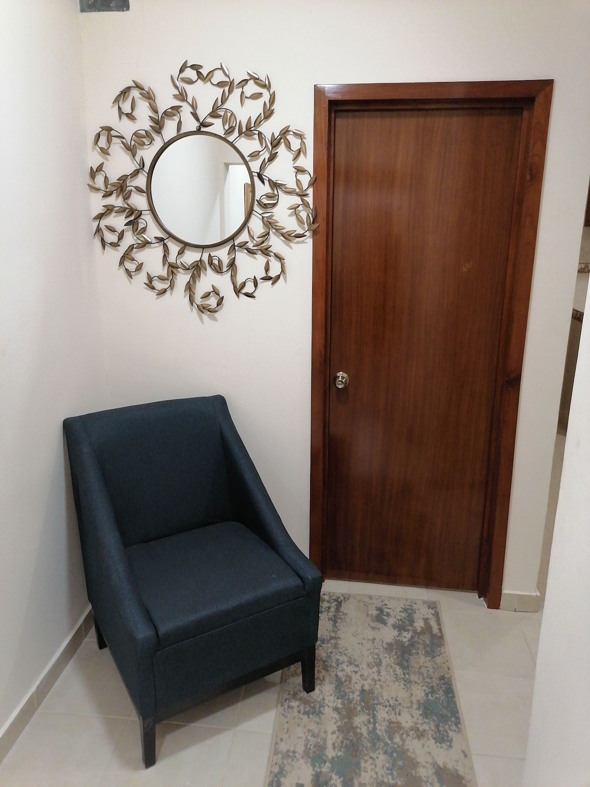A cozy corner features a modern, dark blue accent chair next to a wooden door. Above the chair, a decorative round mirror is surrounded by a simple metallic leaf motif. A neutral-toned area rug lies on the tiled floor, adding texture to the space.