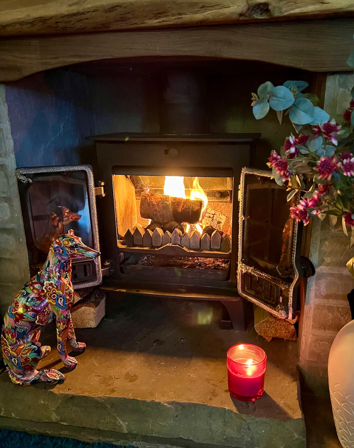 A wood-burning stove is centrally positioned, radiating warmth with a flickering fire inside. A colorful decorative dog statue stands beside it on a stone hearth, while a lit candle glows gently nearby. Fresh flowers are placed in a vase on the right.