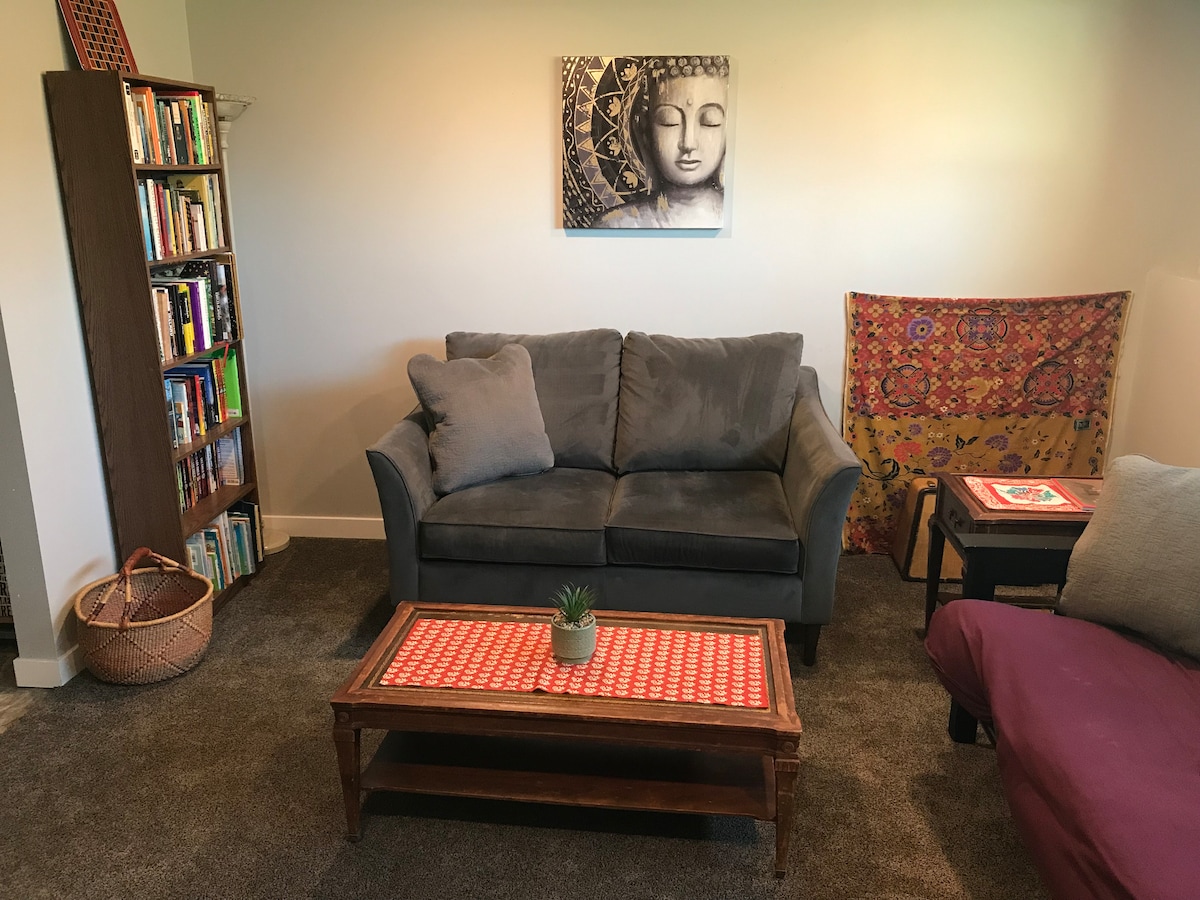 A cozy seating area features a grey sofa with plush pillows and a wooden coffee table, adorned with a plant. Nearby, an eclectic bookshelf displays various books. A decorative fabric hangs on the wall, adding a touch of color to the inviting space.