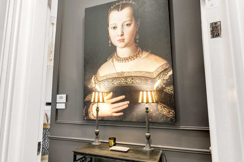 A decorative entryway features a large art piece, partially obscured, highlighting intricate details of the subject's attire. Two elegant table lamps with striped shades provide soft illumination, complementing a console table with a candle and a tablet placed atop.