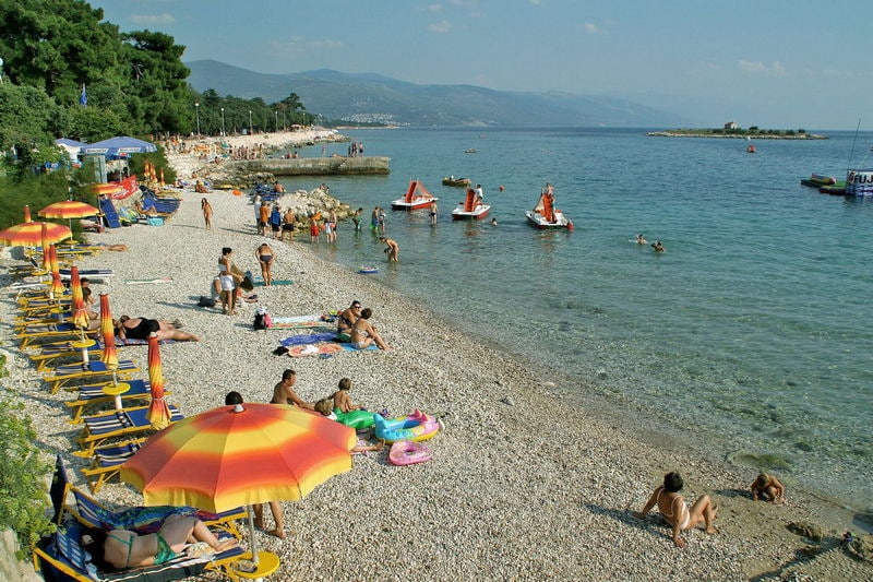 A pebbled beach is filled with sun loungers under colorful umbrellas. People enjoy the water, with some swimming and others on watercraft. Lush trees line the background, and distant hills create a scenic backdrop under a clear blue sky.