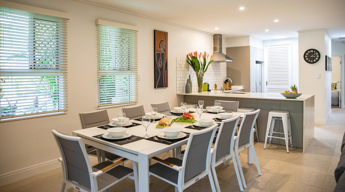 A spacious dining area is highlighted by a large table set for eight, featuring silverware and glassware. Stylish chairs surround the table, while modern kitchen appliances and decor are visible in the background. Natural light filters through window blinds, enhancing the welcoming ambiance.