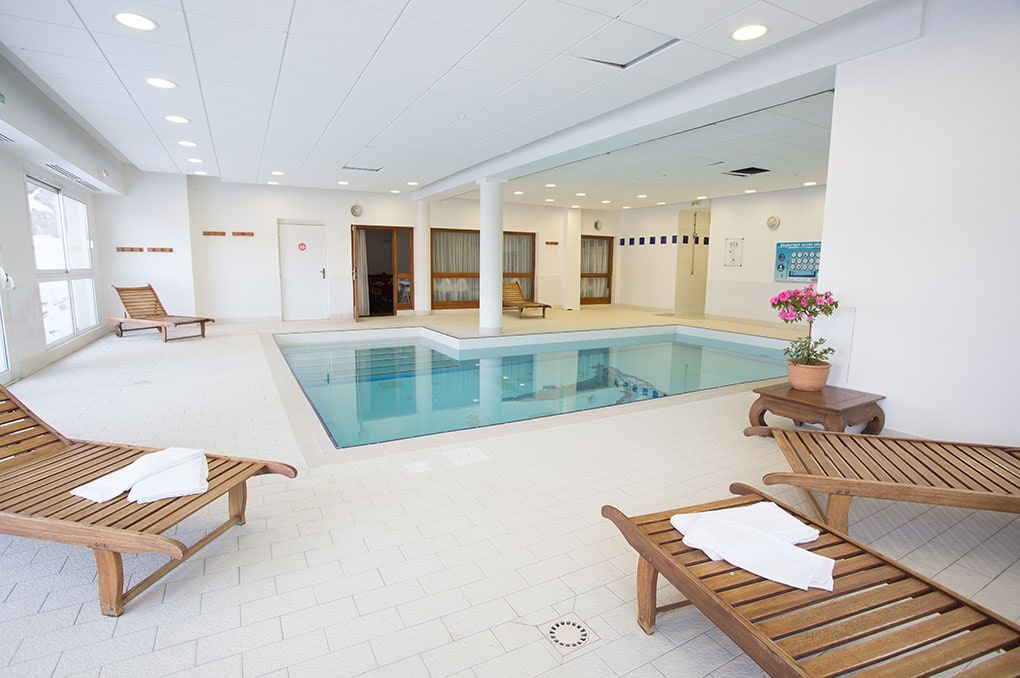 An indoor heated swimming pool area is showcased, featuring a rectangular pool surrounded by light-colored tiles. Several wooden lounge chairs are positioned nearby, with towels neatly folded on each. Soft lighting enhances the serene environment, creating a calm atmosphere.