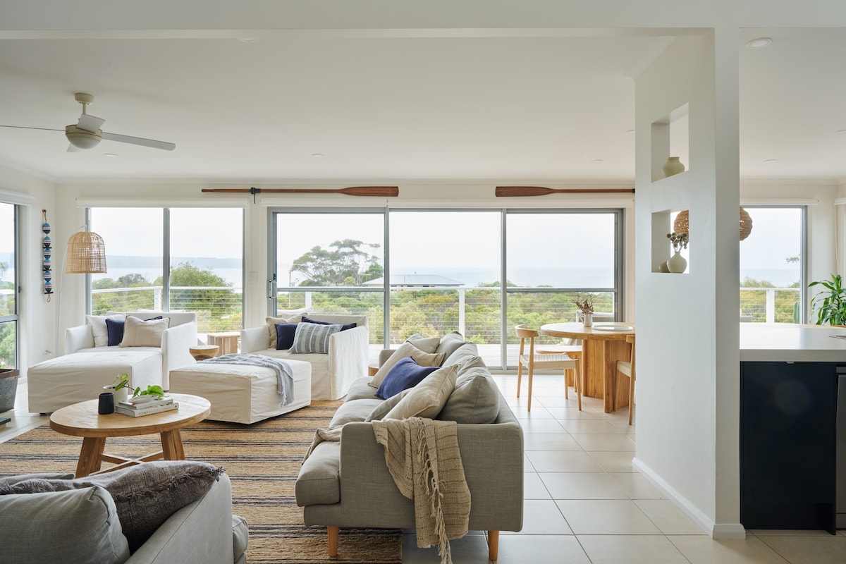 An open-plan living area features large windows overlooking the natural landscape. Two seating areas with light-colored sofas are arranged around wooden tables. A dining area with a round table is visible, enhancing the space's airy feel. Light colors and natural light create a serene ambiance.