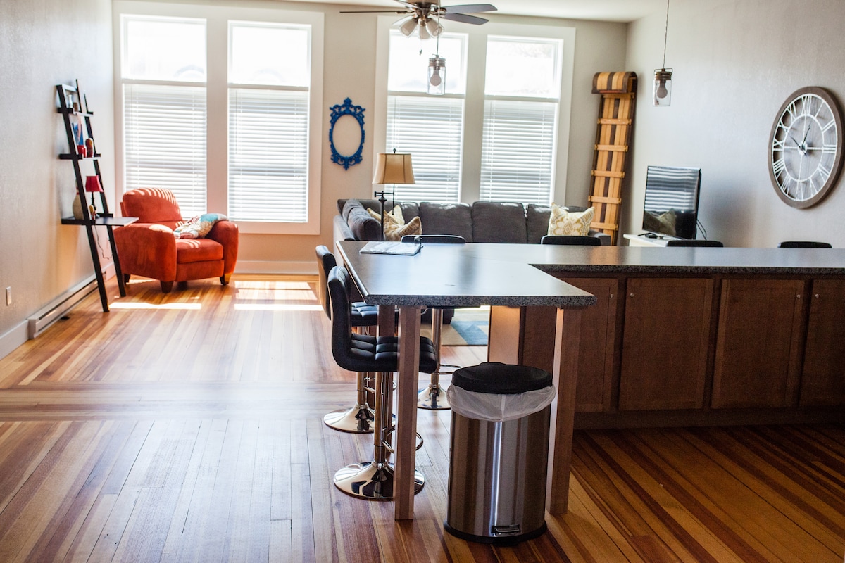An open living area features restored hardwood floors and large windows allowing natural light to fill the space. A cozy red armchair complements a gray sofa. A high-top kitchen bar with stools and a small dining area create a functional layout, with a clock mounted on the wall.