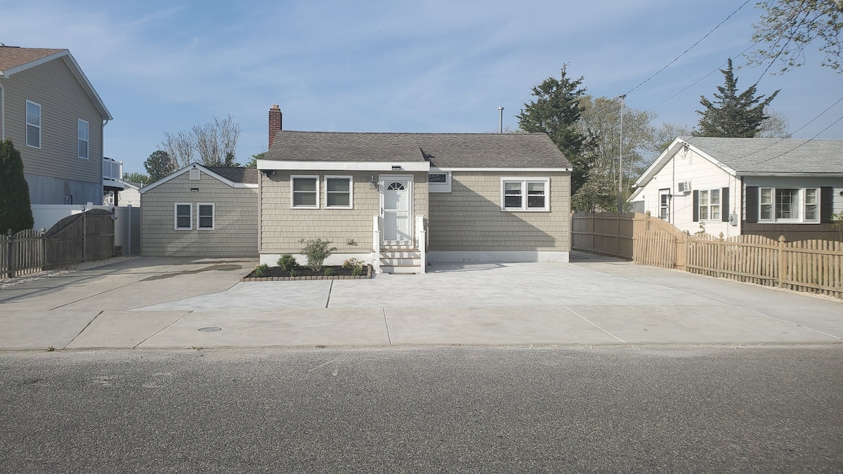 A single-story house is shown with a light brown exterior and white trim. A wide driveway provides ample parking space, surrounded by a well-maintained lawn and flower beds. The property features a welcoming front entrance, with a light-colored door and steps leading to it.