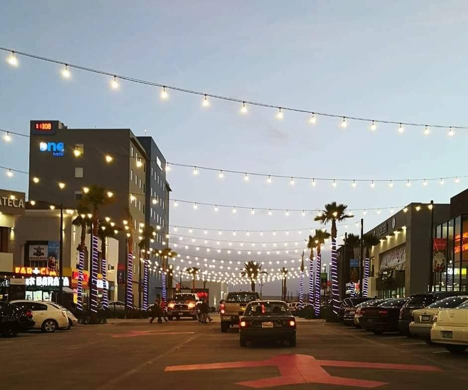 An evening scene showcases a bustling parking area adorned with string lights overhead. Palm trees line the street, and various businesses are visible along the buildings on either side. Vehicles are parked in organized rows, contributing to a lively yet inviting atmosphere.