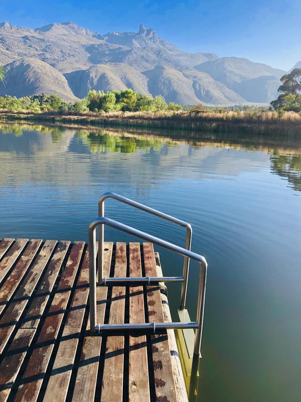 A wooden dock extends into calm waters, with a metal ladder leading down into the lake. Scenic mountains rise in the background, framed by greenery along the shoreline. The reflection of the landscape is visible on the water's surface, enhancing the serene atmosphere.