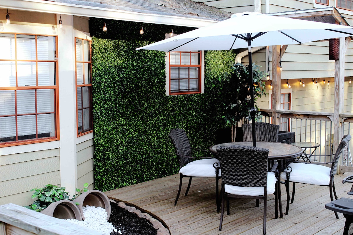 A patio area features a round wooden table surrounded by six chairs, shaded by a large umbrella. A green wall of foliage creates a lush backdrop, while string lights add illumination. Decorative gravel and plants are present in the foreground.