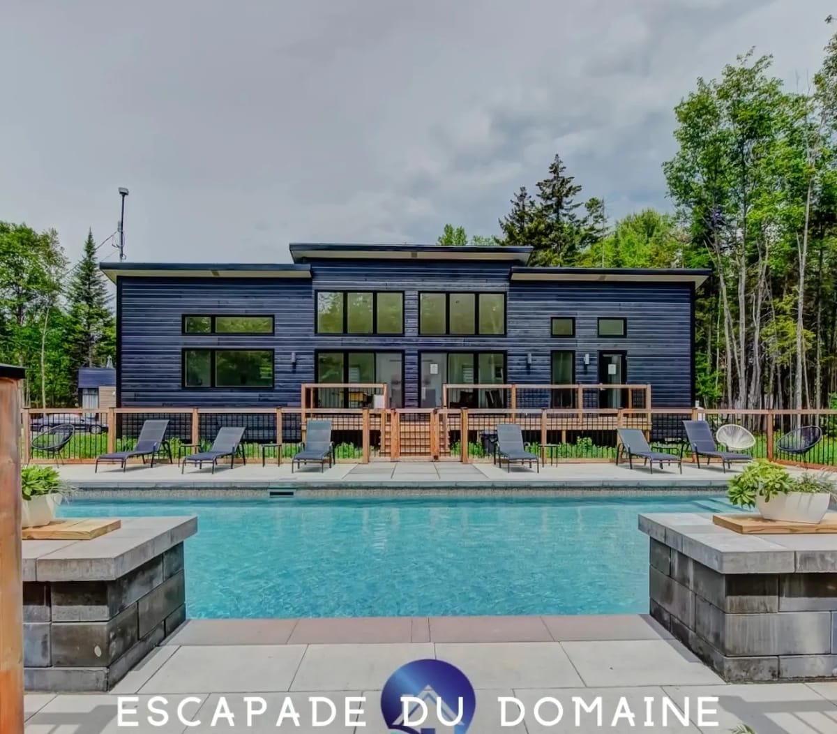 A modern chalet structure with a dark exterior overlooks a spacious pool area. Multiple lounge chairs are arranged around the pool, and large windows are featured throughout. Surrounding greenery adds a natural backdrop, enhancing the tranquil outdoor environment.