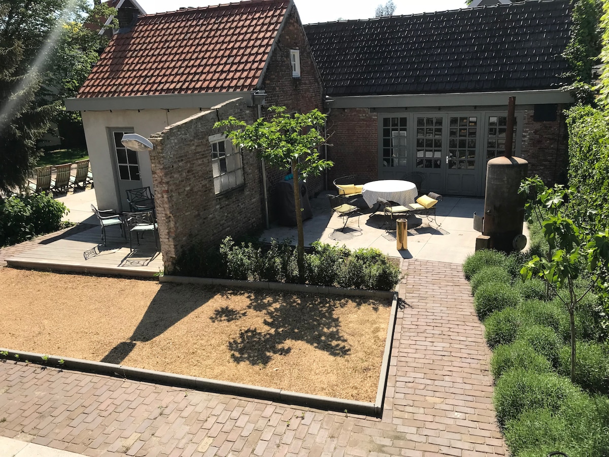 A charming outdoor area is visible, featuring a patio with a round table and chairs. Lush green plants border the space, complemented by a sand-covered section. The rustic brick walls and a traditional roof add character, with a glimpse of a sunny courtyard in the background.