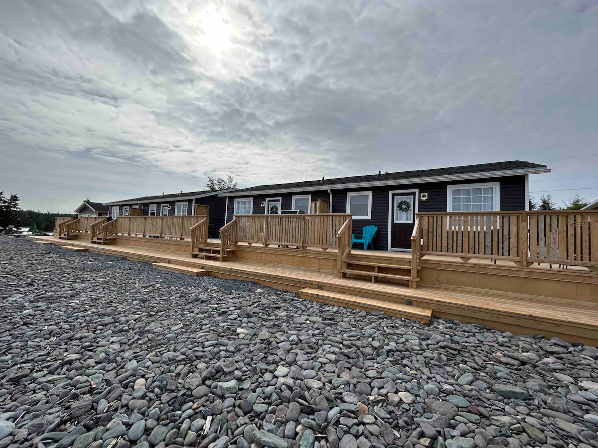 A row of cabin units is positioned along a pebble-strewn coastal area, showcasing wooden decks with railings. Each unit features large windows for natural light, while a blue chair is visible on one of the decks. Overcast skies create a calm atmosphere.