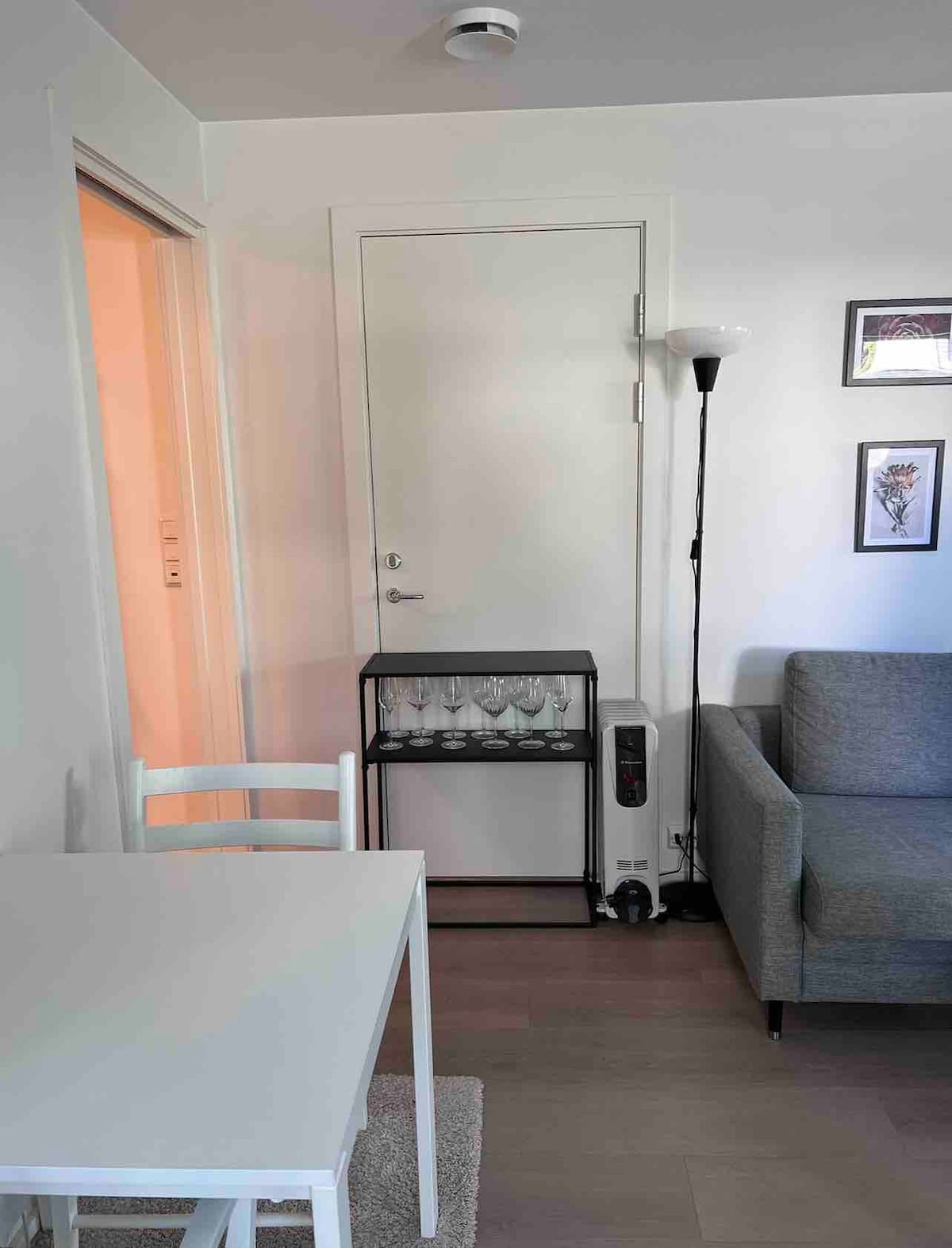 A minimalistic living space features a small dining table with white chairs alongside a stylish gray sofa. A bar cart, holding several glasses, is positioned against a wall near a closed door. Soft lighting from a standing lamp adds a warm touch to the area.