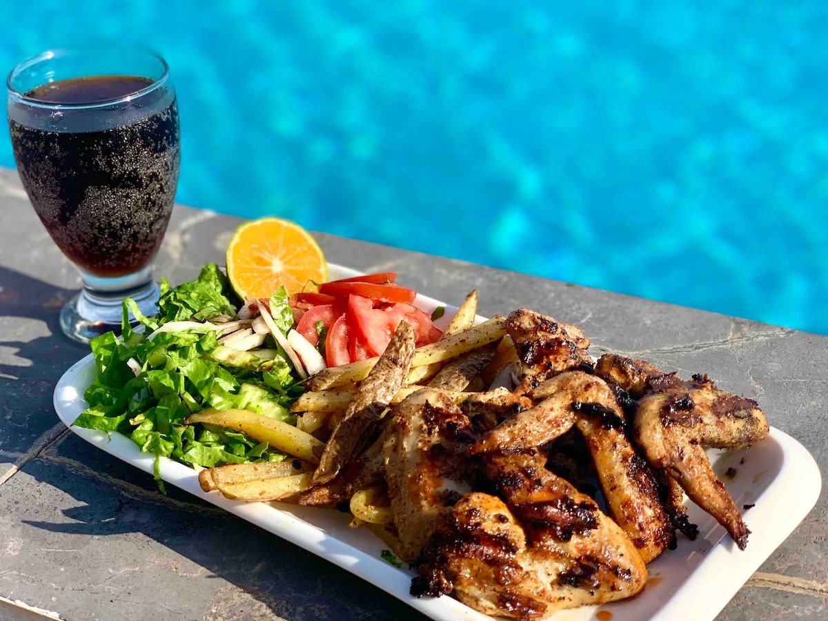 A plate of grilled chicken wings is presented alongside a fresh salad of lettuce and tomatoes, with a serving of French fries. A glass of chilled beverage, garnished with an orange slice, complements the meal. The pool with clear blue water is visible in the background.