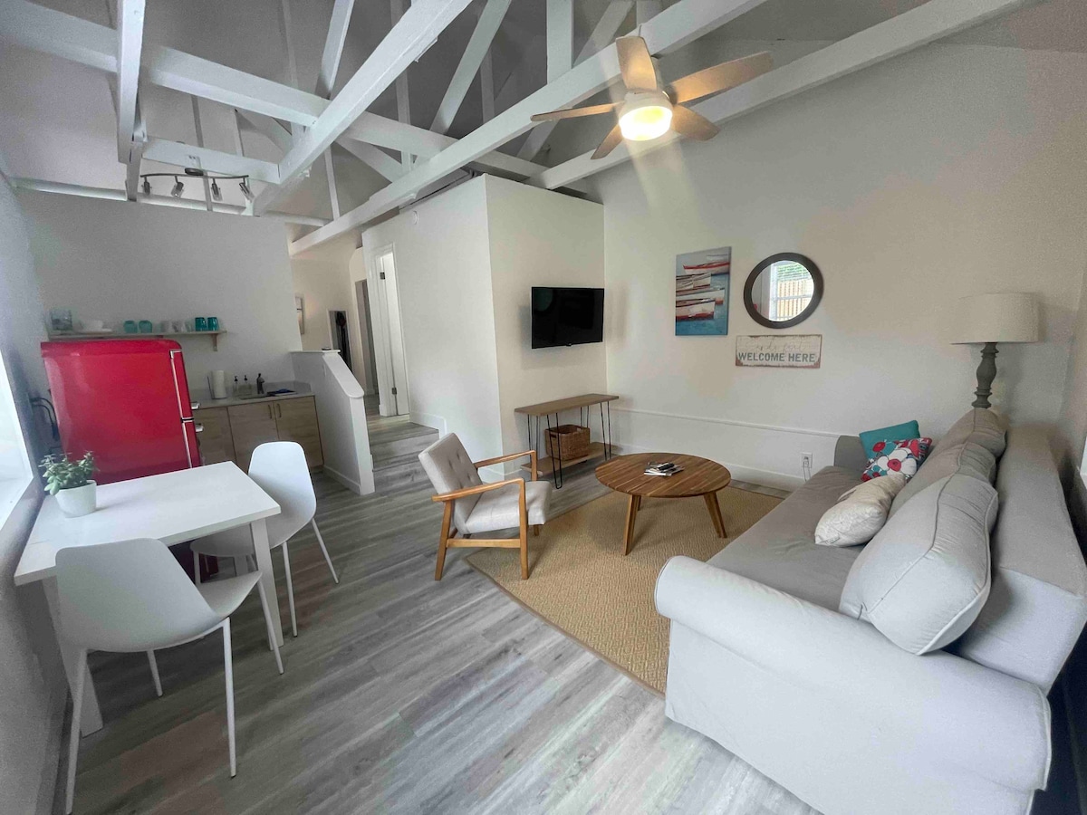 The living area is illuminated by natural light, featuring high, exposed beam ceilings. A comfortable sofa sits beside a round coffee table, and a dining table for four is positioned nearby. A bright red refrigerator adds a pop of color to the clean, modern decor.
