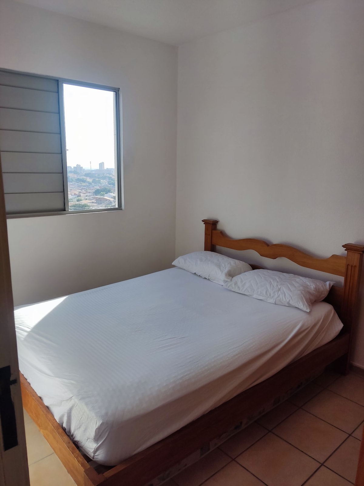A well-lit bedroom features a wooden bed with a soft, white mattress. Natural light enters through a window, illuminating the room. The walls are painted in a light color, creating a calm environment, while a view of the city skyline is visible beyond the window.
