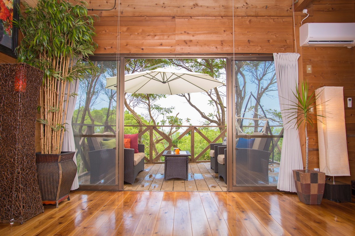 A spacious wooden deck area is depicted, featuring comfortable seating arranged around a small table. Large glass doors lead to a view of lush greenery, and an umbrella provides shade over the outdoor space. Warm wooden tones create a welcoming atmosphere.