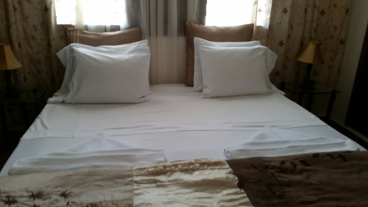 A spacious bed is positioned in the center of the room, adorned with neutral-toned bedding. Two pillows rest at the head of the bed, while decorative towels are neatly arranged at the foot. Natural light enters through two window treatments, illuminating the soft fabric of the curtains.