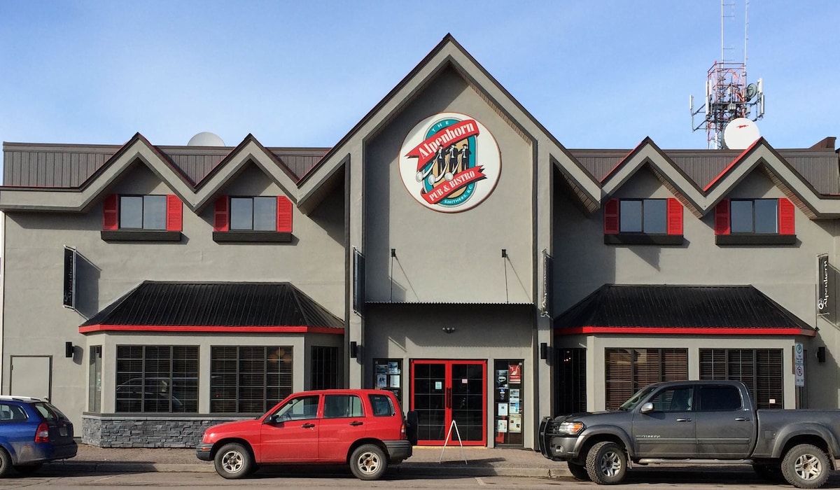 The exterior of the Alpenhorn Bistro & Bar features a modern two-story design with a prominent sign and distinctive triangular rooflines. Large windows allow natural light to fill the space, and a mix of vehicles is parked along the street, reflecting the lively downtown atmosphere.