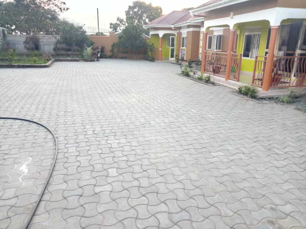 A spacious and well-maintained outdoor area features a patterned paved surface, surrounded by greenery and plants. Nearby, colorful exterior walls of the adjacent buildings create a welcoming atmosphere with open access to front porches.