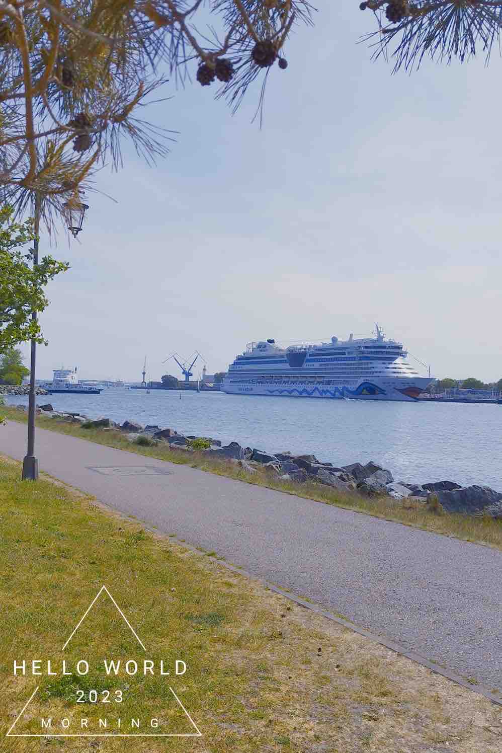 A serene waterfront scene is depicted, featuring a large cruise ship docked near the shore. Lush green grass borders a paved path, which runs parallel to the water, while a clear sky provides a bright backdrop for the bustling maritime activity.