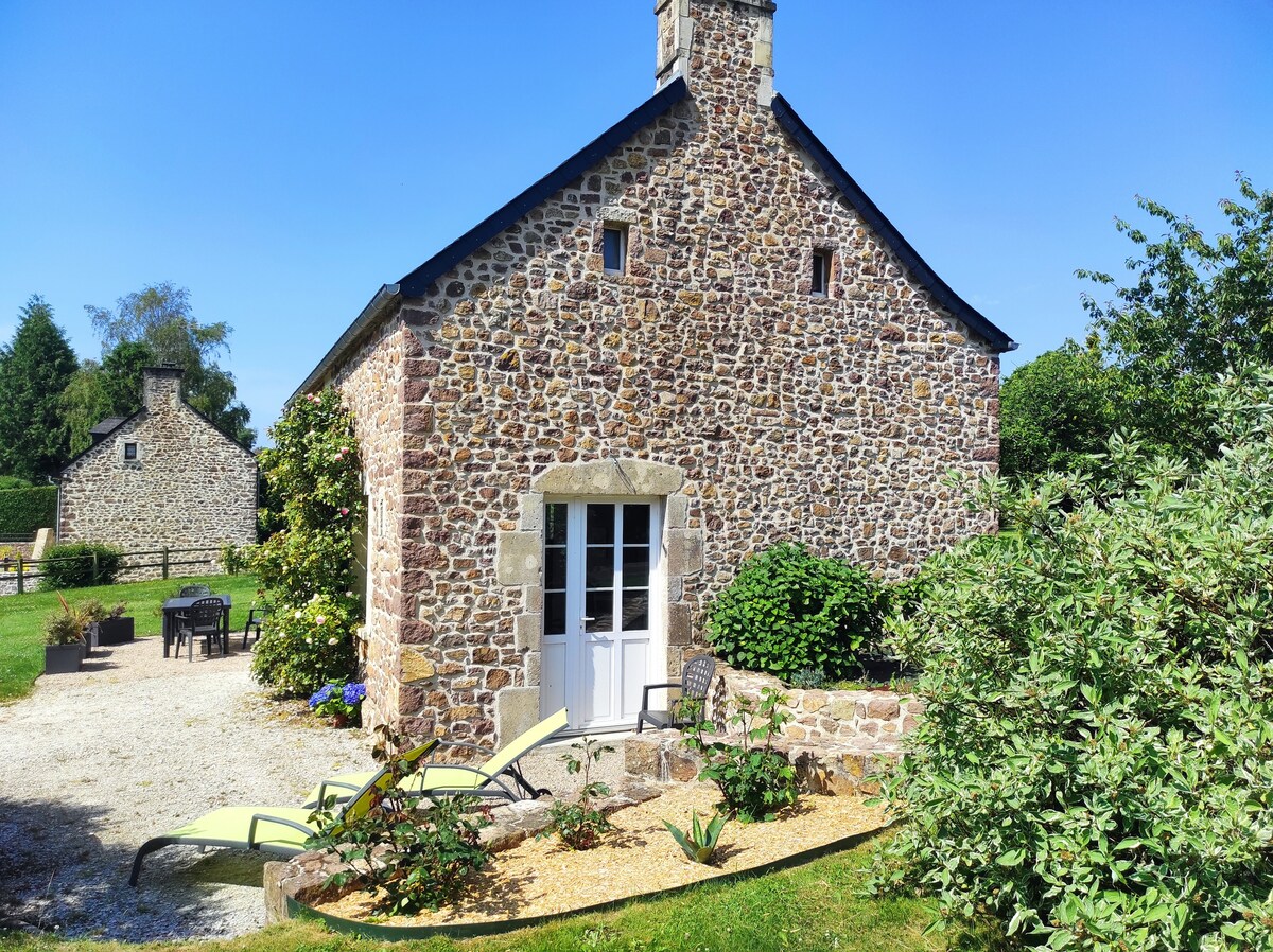 An exterior view of a beautifully renovated stone cottage is displayed, surrounded by a lush garden. Sun loungers and a small seating area are positioned on a pebbled patio, while a clear blue sky enhances the serene atmosphere.