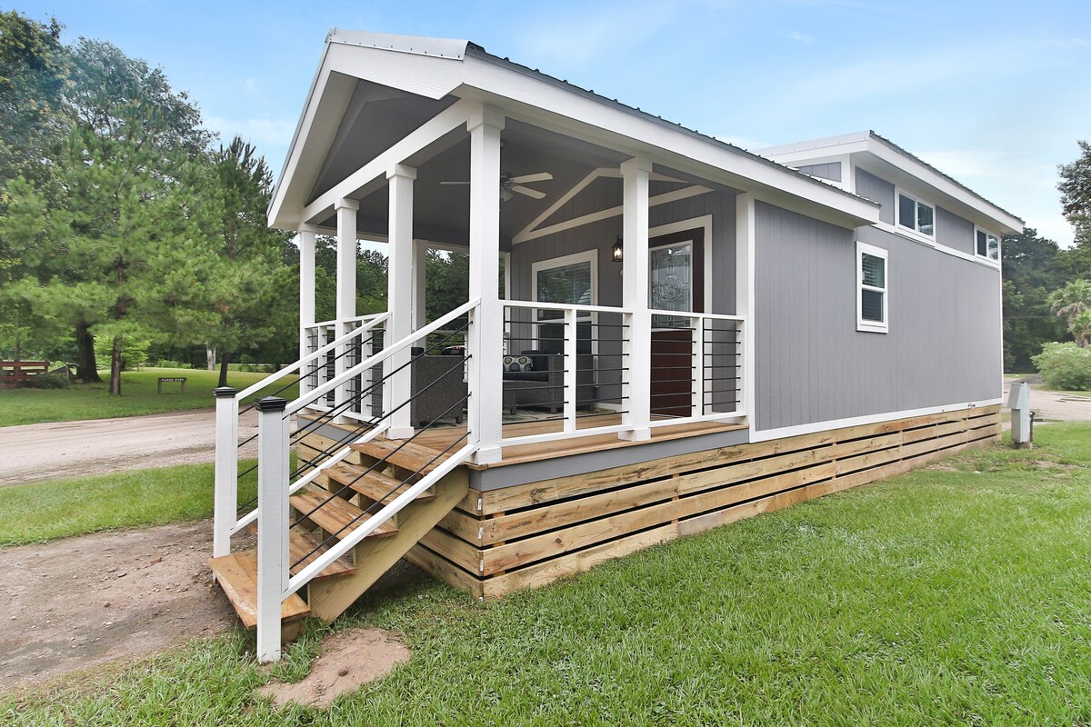 The exterior of the tiny house features a welcoming front porch with a railing and steps leading up. Grass surrounds the area, and trees can be seen in the background, enhancing the natural setting of the home.