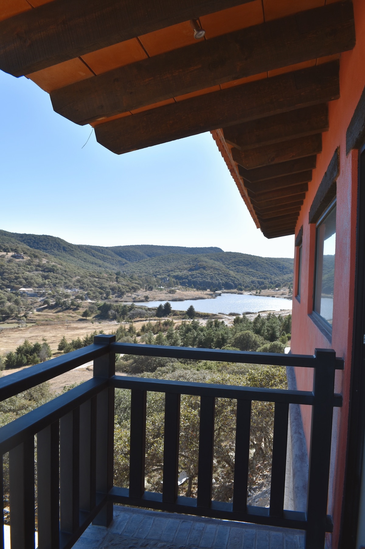 A view from a balcony showcases expansive hills and a serene body of water below. The surrounding landscape features lush greenery and gentle slopes, framed by a wooden railing and part of the villa's exterior.