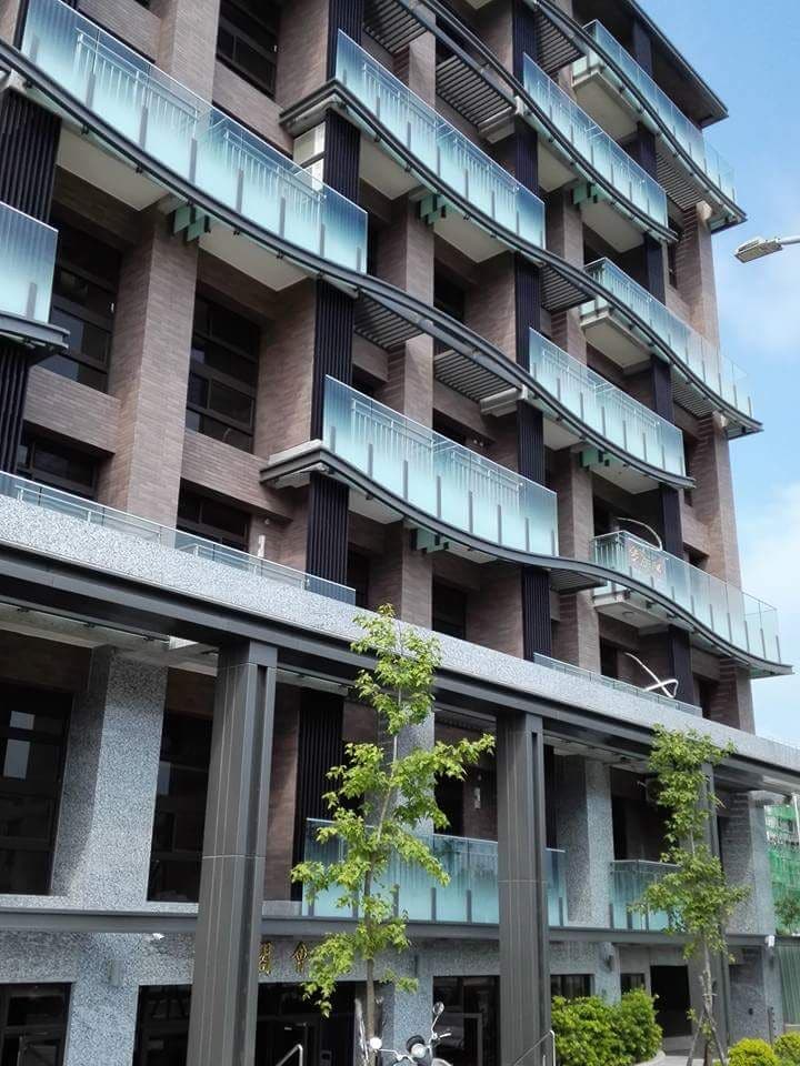 The exterior of a modern apartment building is showcased, featuring multiple floors with glass balconies and a contemporary façade. Trees line the entrance, and the building's design is characterized by a combination of stone and glass materials.