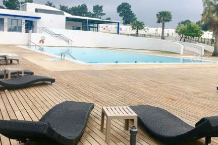 An outdoor area is presented with a wooden deck surrounding a swimming pool. Lounge chairs are arranged neatly alongside the pool, with a small table placed nearby. The facility is bordered by green landscape and a building is visible in the background.