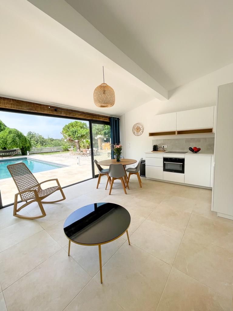 A bright living space features a modern kitchen and a small dining area with wooden chairs around a round table. Large glass doors open to reveal a sunny terrace and pool view, enhancing the area with natural light.